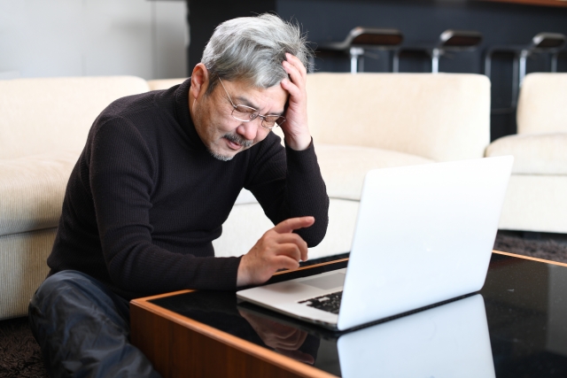黒いシャツを着たおじいちゃんがパソコンの前で手を頭にあてて悩んでいる