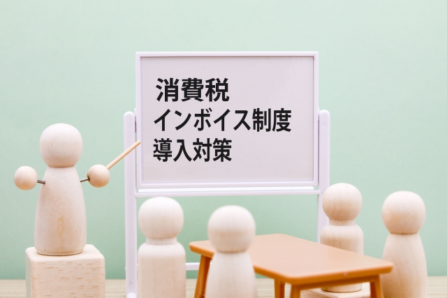 ホワイトボードに消費税、インボイス制度、導入対策という文字が書かれていて、人間の形をした模型の4体が机の周りに、1体が棒をもって説明している